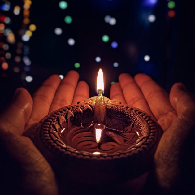 A lit candle in a diya, held in 2 hands