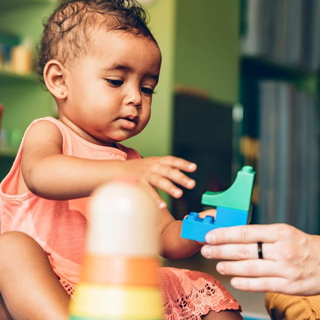 a toddler in a pink dress is reaching for a toy held by an adult