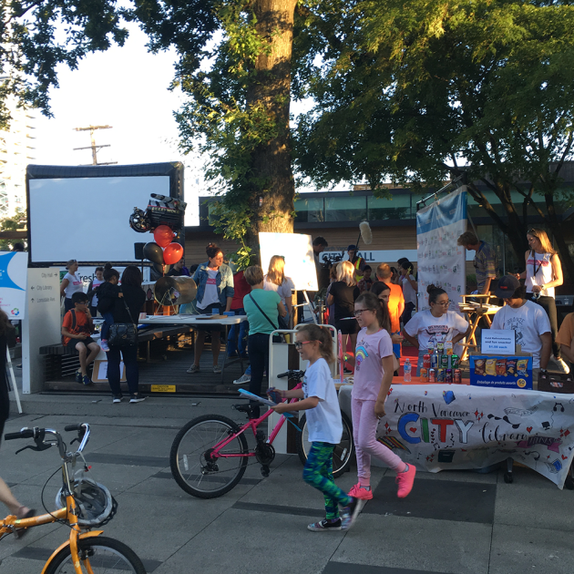 a movie night in the plaza with a large screen in the background and teens selling popcorn and chips at a table in the foreground