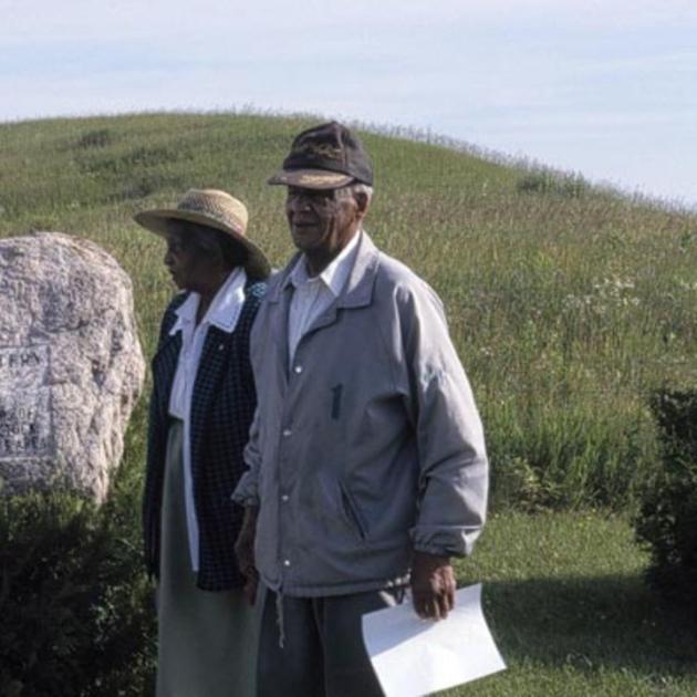 An elderly Black man and woman stand next to a large stone tombstone in a grassy field