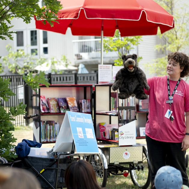 A curly haired person in a red shirt, animating a bear puppet for the audience. They're standing in front of the open book bike library.