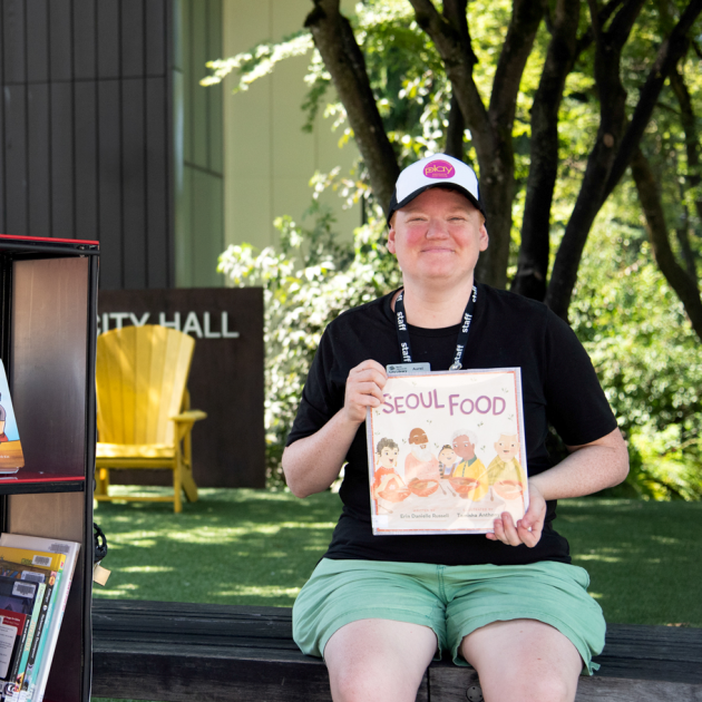 A person in a baseball cap and black t-shirt sitting next to the library's book bike and holding up a book