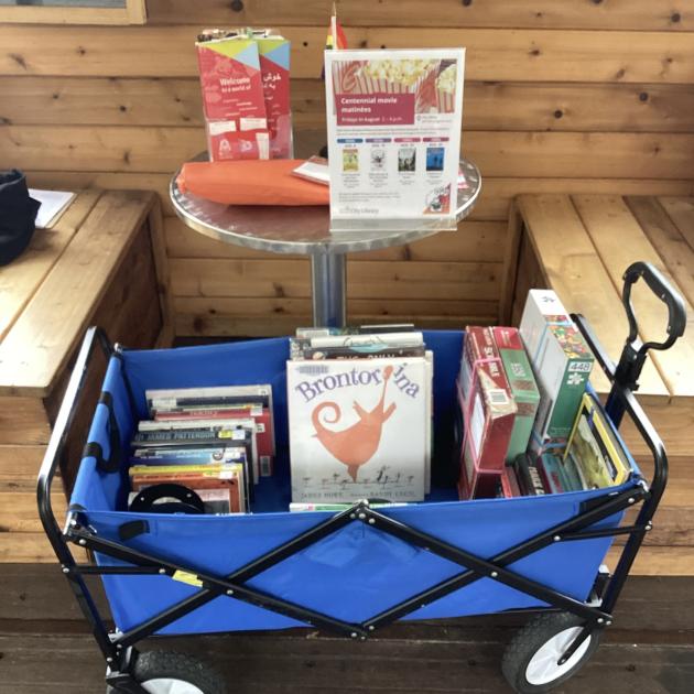 A blue wagon full of books for all ages is set up in a city parklet. On the table behind it, there's a poster promoting a library event and general information.