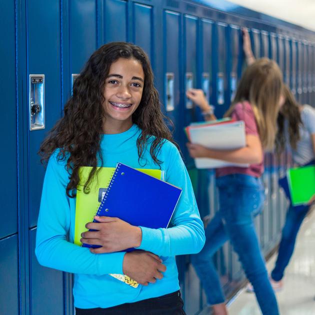 Teen in bright blue shirt leaning against blue high school lockers, holding binders and workbooks