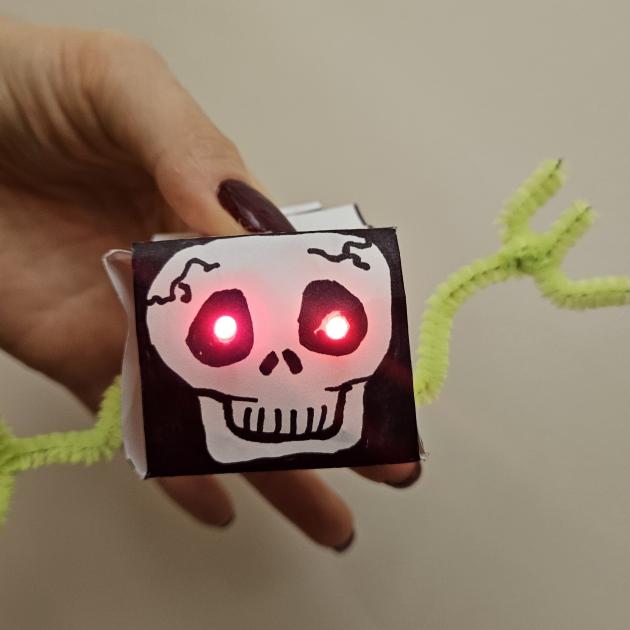 3D paper circuit is a cube box made from paper. It has a scull face drawn on it with red LED light up eyes and neon yellow pipe-cleaner arms. Someone holds it so the eyes light up. . 