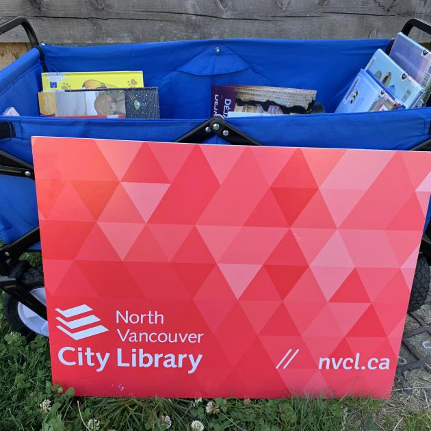 A blue wagon loaded up with books. In front of it, a giant version of the North Van City Library card.