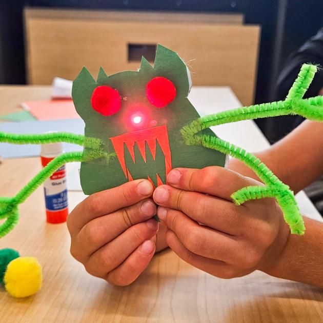 Child's hands holding paper monster with green pipe cleaners and light-up nose