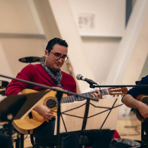 A man playing a guitar wearing spectacles, a red sweater and patterned scarf.