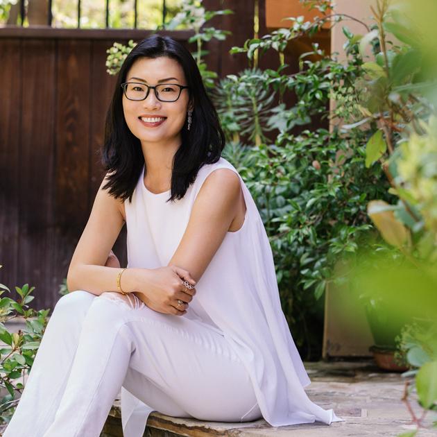 A photograph of YA author Kelly Yang. She is dressed in all white with green foliage around her.