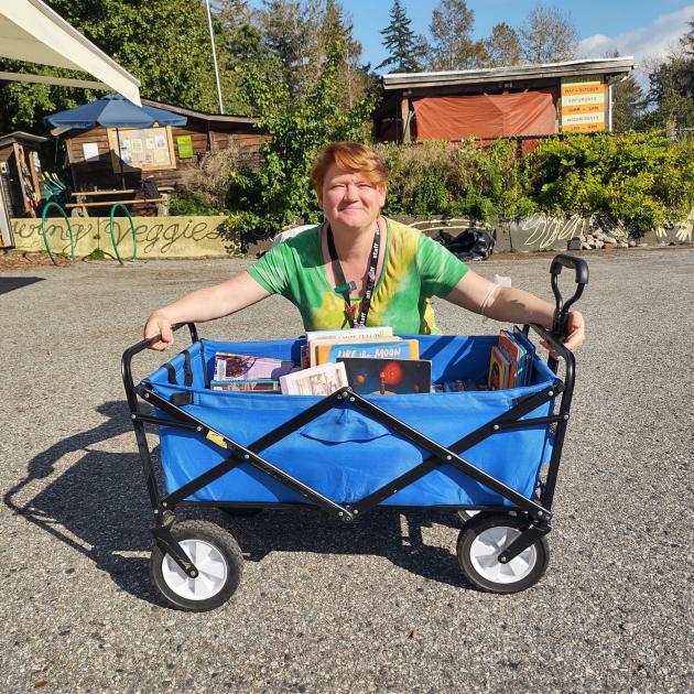 A small blue portable 'truck' containing books, with a staff member crouching behind it and smiling