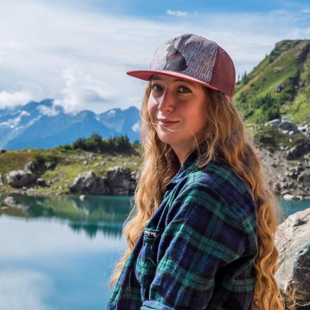 Lianne Oelke, author, with long wavy blonde hair and a blue and green plaid shirt, smiles at the camera from beneath a trucker hat. In the background there are the green slopes and blue of the mountains and a mountain lake.