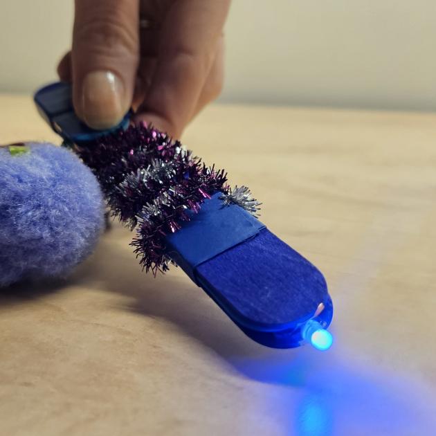 A DIY light-up wand. Wand is made of blue popsicle sticks and have purple and silver pipe-cleaners wrapped around the stem. There is a purple pompom attached the the back. You can see a hand holding the back of the wand and pressing a button to turn on the blue LED light at the front end.