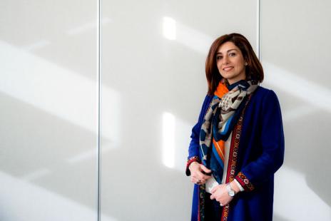 Portrait of a woman wearing a colorful scarf and blue sweater standing in front of a plain grey wall